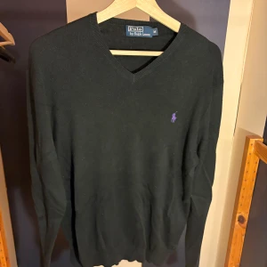 Svart v-ringad tröja Polo Ralph Lauren - Svart v-ringad tröja från Polo Ralph Lauren med lila broderad logga på bröstet. Tröjan är långärmad och har en klassisk, stilren look. Perfekt för dig som gillar enkel och tidlös stil.