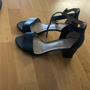 Svarta sandaler med klack från Clarks - Stilrena svarta sandaler från Clarks med bred rem över tårna och justerbart spänne vid ankeln. Skorna har öppen tå och en stabil blockklack som ger extra höjd. Tillverkade i skinn med en klassisk och tidlös design som passar till många outfits.