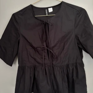Svart blus med knyt från H&M Divided - Svart blus från H&M Divided i storlek XS. Modellen har knyt framtill och en lätt volang nedtill. Blusen har korta ärmar och rund halsringning, perfekt för en trendig och avslappnad look.