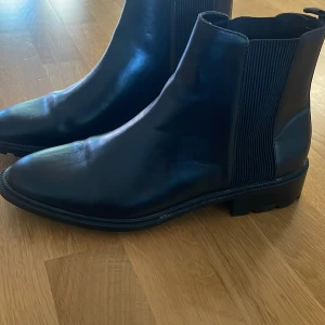 Svarta chelsea boots i skinn - Stilrena svarta chelsea boots med spetsig tå och platt sula. Skorna är tillverkade i slätt skinn och har elastiska paneler på sidorna för enkel påtagning. Perfekta för dig som gillar en klassisk och clean look.