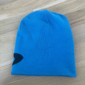  mössa från Oakley blå/vit - Snygg mössa från Oakley i 100% akryl. Ena sidan är klarblå med svart Oakley-logga, andra sidan är vit med blå kant och broderad Oakley-text. 