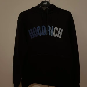 Svart hoodie från Hoodrich med blå text - Svart hoodie från Hoodrich i storlek M med broderad text i blå och vita nyanser över bröstet. Klassisk huva med snörning och känguruficka. Bra skick och en snygg och varm tröja perfekt nu till vintern.