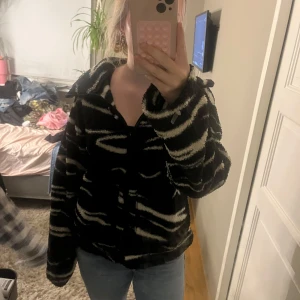 Svart teddyjacka med zebramönster - Säljer en svart teddyjacka med vitt zebramönster och dragkedja framtill. Jackan är fluffig och har en avslappnad passform med hög krage och långa ärmar. Perfekt för dig som vill sticka ut med en cool och mysig look.