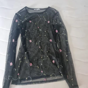 Svart meshblus med broderade blommor - Säljer en transparent svart meshblus från Primark med broderade blommor i rosa och grönt. Blusen har lång ärm och en smal passform, perfekt för att styla lager på lager eller med ett linne under. Superfin detalj med de små blommorna som ger en romantisk vibe.