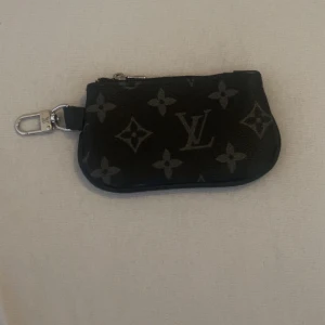  svart monogram plånbok - Snygg liten plånbok i svart skinn med klassiskt monogrammönster och silverfärgad dragkedja. Perfekt att haka fast i väskan eller nyckelknippan med den smidiga karbinhaken. Stilren och ikonisk accessoar för dig som gillar lyxiga detaljer.