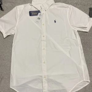 Vit kortärmad skjorta Polo Ralph Lauren - Klassisk vit skjorta från Polo Ralph Lauren med korta ärmar och button-down krage. Skjortan har broderad blå logga på bröstet och stängs med vita knappar framtill. Tillverkad i mjuk bomull, perfekt för en clean och stilren look.