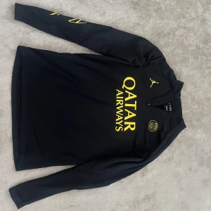 Svart PSG långärmad fotbollströja - Snygg svart långärmad PSG-tröja från Jordan med gul logga och Qatar Airways-tryck på bröstet. Tröjan har halv dragkedja framtill och är tillverkad i ett lätt och stretchigt funktionsmaterial som passar perfekt till träning eller match.