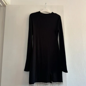 Svart ribbad klänning från Zara - Säljer en svart ribbad kortklänning från Zara med långa ärmar och rund hals. Klänningen har snygga fransar längst ner som ger en cool touch. Perfekt för dig som gillar stilrena och trendiga plagg.