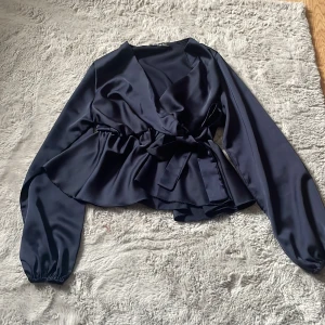 Mörkblå omlottblus i satin från Missäy - Säljer en mörkblå omlottblus från Missäy i glansig satin. Blusen har långa ärmar med resår vid handlederna, knytband i midjan och en snygg v-ringning. Perfekt för dig som gillar en chic och trendig look.