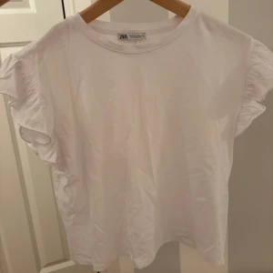 Vit t-shirt med volangärmar från Zara - Nyskick!! Vit t-shirt från Zara med korta ärmar och volangdetaljer på axlarna. Rund halsringning och loose fit.
