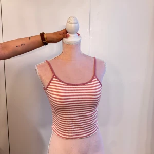 Randig croppad linne i rosa och vit - Superfin croppad linne med tunna axelband och rosa-vita ränder. Perfekt för varma dagar och ger en chill vibe. Ribbad känsla i materialet och smickrande passform som sitter snyggt på kroppen.