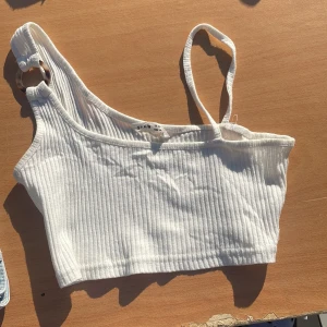 Vit ribbad oneshoulder topp från SHEIN - Snygg vit ribbad topp från SHEIN med oneshoulder-design och en cool ringdetalj på axelbandet. Toppen är croppad och har smala axelband, perfekt för en trendig look. Materialet är mjukt och stretchigt, vilket gör den superbekväm.