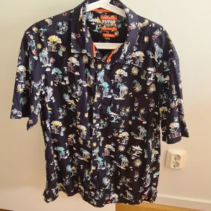 Svart mönstrad skjorta från Superdry - Säljer en svart skjorta från Superdry med tropiskt mönster i gult, turkost, vitt och orange. Skjortan har korta ärmar, klassisk krage och knäppning framtill. Perfekt för dig som vill sticka ut med en färgglad vibe. Materialet känns lätt och skönt mot huden.