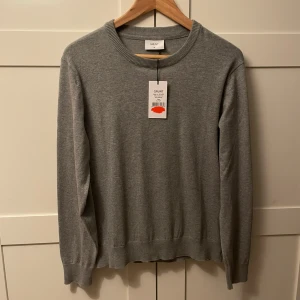 Grå stickad tröja från Grunt XL - En stilren grå stickad tröja från Grunt i storlek XL i barn. Tröjan har rund halsringning, ribbade muddar och är långärmad. Perfekt för lager-på-lager och funkar till många olika outfits. Helt ny med prislapp.