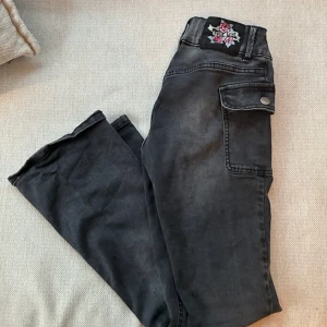 Svarta cargopants från Lindex, stl 152 - Snygga svarta cargopants från Lindex med raka ben och coola fickor med lock på sidorna. Byxorna har en broderad detalj bak i midjan och dubbla knappar framtill. Perfekta för en avslappnad och trendig look!❤️❤️