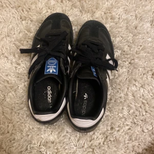 Adidas Samba svarta sneakers - Klassiska Adidas Samba sneakers i svart skinn med vita detaljer och de ikoniska tre ränderna på sidorna. Skorna har bruna gummisulor, svart snörning och blå Adidas-logga på plösen. Perfekta för en sportig och stilren look.
