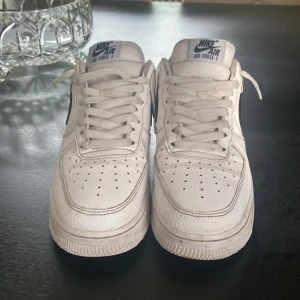 Nike Air Force 1 vita och svarta sneakers - Klassiska Nike Air Force 1 sneakers i vitt skinn med svarta detaljer och svart swoosh. Lågt skaft, snörning och perforerad tå för extra ventilation. Perfekta för dig som gillar en clean och tidlös streetstil.Jag säljer dessa för att de har blivit för små för mig 