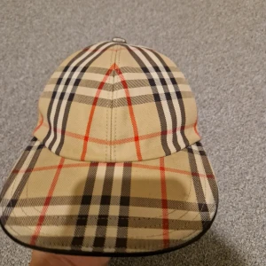 Burberry rutig keps i beige och svart - Snygg Burberry keps med klassiskt rutigt mönster i beige, svart och rött. Modellen är en trucker cap med böjd skärm och justerbar passform. Tillverkad i bomull och har märkets ikoniska design som ger en lyxig vibe till din outfit.