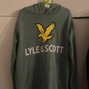 Grön hoodie från Lyle & Scott Junior - Snygg grön hoodie från Lyle & Scott Junior med stor gul fågel-logga och vit text på bröstet. Tröjan har huva, känguruficka och långa ärmar. Perfekt för dig som gillar streetwear och vill sticka ut med en klassisk look.