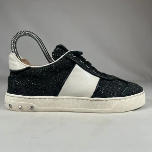 Valentino Garavani svarta sneakers med glitter - Svarta och vita Valentino Garavani sneakers med glittrig ovandel och vita skinnpaneler. Klassisk rund tå, snörning och ikoniska nitar på sulan. Snygg kontrast mellan svart glitter och vitt skinn, perfekt för dig som vill sticka ut.