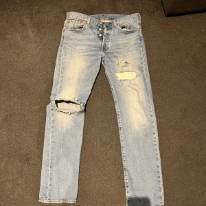 Levi's 501 ljusblå slitna jeans - Ljusblå Levi's 501 jeans med klassisk knappgylf och raka ben. Jeansen har slitningar och hål på knäet och låret för en cool, avslappnad look. Tillverkade i jeansmaterial med vintagekänsla och ikoniska bakfickor med röd Levi's-tagg.