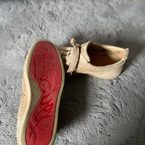 Beige mockasneakers från Louboutin - Snygga sneakers från Christian Louboutin i beige mocka med klassisk röd sula och snörning. Skorna har rund tå och platt sula, samt diskreta ventilationshål på sidorna. Perfekta för dig som vill sticka ut med exklusiva detaljer. Defekt finns som syns på bild 3 annars är dom fantastiska🤩