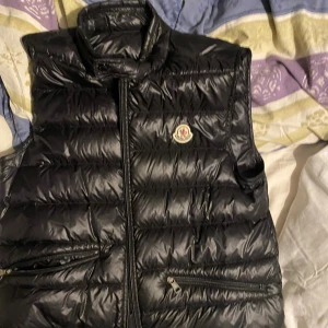 Svart dunväst från Moncler - Säljer en svart, glansig dunväst från Moncler med hög krage och två dragkedjefickor framtill. Västen har klassisk quiltad design och ikonisk Moncler-logga på bröstet. Perfekt för lager-på-lager och riktigt snygg till streetwear eller casual outfits.