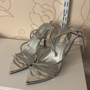 Silvriga klackar med strass från Nelly - Glittriga silvriga pumps med smala remmar täckta av strass och spetsig tå. Skorna har hög klack och elegant design med flera remmar som slingrar sig runt foten och ankeln. Perfekta för att sticka ut och ge extra bling till din outfit.