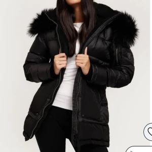 Svart pufferjacka med fuskpäls från Rockandblue - Snygg svart pufferjacka från Rockandblue med stor huva och fuskpäls. Köpt för 4500kr och har använts i cirka två veckor. Jackan har dragkedja, flera fickor och justerbar midja. Perfekt för kalla dagar med sin vadderade design och coola detaljer. Passar dig som vill ha en varm och stilren vinterjacka. 