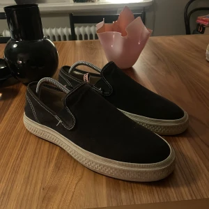 Svarta slip-on sneakers från Acne Studios - Svarta slip-on sneakers från Acne Studios i canvas med vita kontrastsömmar och beige sula med unikt mönster. Klassisk rund tå och platt sula, perfekt för en clean och stilren look. Diskret Acne Studios-logga på insidan. Strl 44, 1400kr!