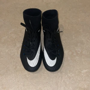 Nike Hypervenom fotbollsskor svart/vit - Säljer ett par Nike Hypervenom fotbollsskor i svart med vita detaljer och stora Nike-loggor. Skorna har snörning, hög strumpliknande krage och metall-dobbar för bästa grepp på planen. Tillverkade i syntetmaterial med textur för bättre bollkontroll.