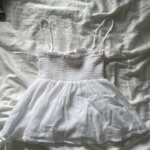 Vit smockad linnetopp Gina Tricot - Supersöt vit linnetopp från Gina Tricot med smockad överdel och tunna axelband. Toppen har en luftig, lätt utställd nederdel och volangkant upptill. Perfekt för varma dagar och enkel att matcha med jeans eller kjol. Endast testad!