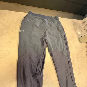 Under Armour SpeedPocket träningsbyxor M - Säljer ett par mörkgrå Under Armour SpeedPocket träningsbyxor i storlek M. Byxorna har slim passform, elastisk midja och är gjorda i syntetmaterial som andas. Perfekta för löpning eller gym, med praktisk ficka för mobilen. Snyggt logotryck på benet.