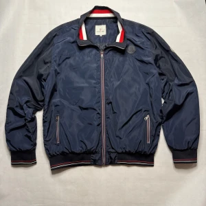 Marinblå vindjacka från Moncler - Snygg marinblå vindjacka från Moncler med klassiska ränder i rött, vitt och blått på krage och muddar. Jackan har dragkedja framtill, två fickor med dragkedja och Moncler-logga på bröstet och ärmen. Perfekt för dig som vill ha en stilren och sportig look.  STORLEK 4XL
