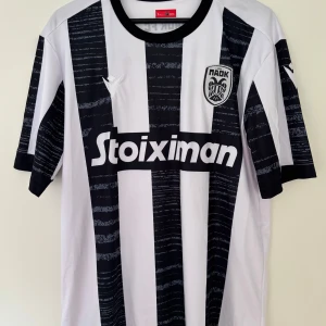 PAOK FC randig fotbollströja Macron - Snygg PAOK FC matchtröja från Macron med breda svarta och vita ränder och coolt tryck på bröstet. Kortärmad modell med rund hals och Stoiximan sponsortryck framtill. Materialet är lätt och andas, perfekt för träning eller match.