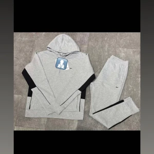 Grå hoodie från Lacoste med svarta detaljer - Snygg grå hoodie från Lacoste med svarta paneler på sidorna och ärmarna. Klassisk huva och diskret Lacoste-logga på bröstet. Perfekt för en chill och sportig look. Mjuk bomullskänsla och långärmad modell.