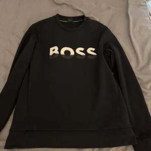 Svart tröja från Boss - Säljer en stilren svart tröja från Boss med deras logga i vitt på bröstet. Tröjan har en klassisk rund hals och långa ärmar. 