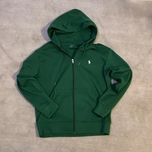 Ralph Lauren hoodie grön - Säljer en snygg grön hoodie från Ralph Lauren med dragkedja och klassisk logga på bröstet. Tröjan har långa ärmar och en praktisk huva med snörning. Perfekt för en avslappnad stil. Köpt på vinted.