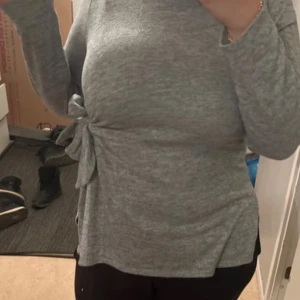 Grå tröja med knytning - Säljer en stilren grå tröja med knytning i sidan. Tröjan har långa ärmar och en bekväm passform, perfekt för både vardag och lite finare tillfällen. Den är tillverkad i ett mjukt material som känns skönt mot huden. Den passar dig som har storlek L/M