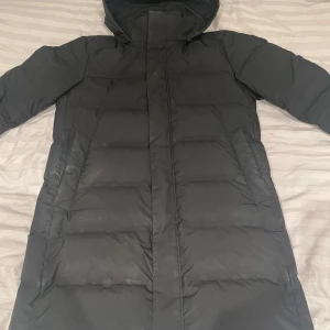Svart dunjacka från Uniqlo - Säljer en svart lång dunjacka från Uniqlo med huva och dragkedja. Jackan har långa ärmar och är perfekt för kalla dagar. Den är stilren och funktionell med en klassisk design.