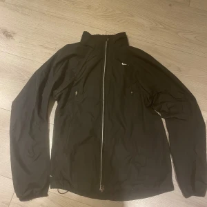 Svart vindjacka med dragkedja - Säljer en stilren svart vindjacka med dragkedja framtill och två sidofickor med dragkedjor. Jackan har en hög krage och är perfekt för blåsiga dagar. Den har en liten logga på bröstet och en praktisk ficka med dragkedja på ryggen.