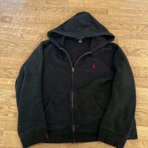 Svart hoodie från Ralph Lauren - Säljer en klassisk svart hoodie från Ralph Lauren med dragkedja och huva. Den har en röd broderad logotyp på bröstet och är tillverkad i ett mjukt material. Perfekt för en avslappnad stil. Storlek 14-16 men skulle säga Xs i vuxen storlek 