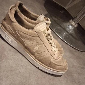Beige sneakers från Polo Ralph Lauren - Snygga beige sneakers från Polo Ralph Lauren i mocka. Skorna har en klassisk design med snörning och en diskret logga på sidan. Perfekta för en stilren look.