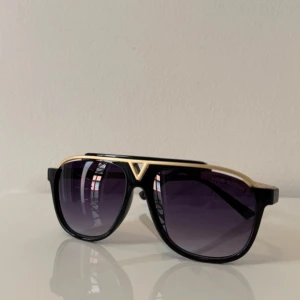 Louis Vuitton - Snygga svarta solglasögon från Louis Vuitton med gulddetaljer och mörka linser. Den ikoniska V-formen i fronten ger en lyxig touch. Perfekta för att höja din stil till nästa nivå.