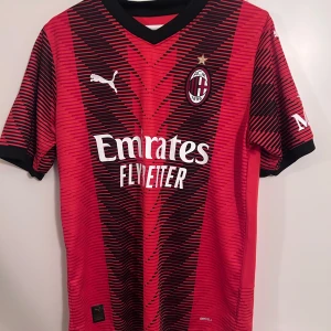 Röd och svart AC Milan fotbollströja från Puma - Säljer en snygg röd och svart AC Milan fotbollströja från Puma. Tröjan har korta ärmar och ett randigt mönster. Den har klubbens emblem på bröstet och sponsorer tryckta på framsidan och baksidan. Perfekt för alla AC Milan-fans!