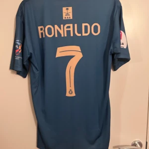 Ronaldo Tröja - Säljer en blå fotbollströja från Nike med korta ärmar. Tröjan har ett tryck med texten 'KAFD' på framsidan och 'Ronaldo 7' på baksidan. Perfekt för fotbollsfans som vill visa sitt stöd.