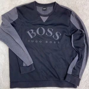 Svart och grå tröja från Hugo Boss - Snygg svart tröja från Hugo Boss med gråa ärmar och logotyp framtill. Perfekt för en stilren look.