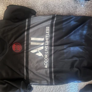 Svart fotbollströja från Nike - Snygg svart fotbollströja från Nike med klubbmärke på bröstet och sponsorlogga i vitt. Tröjan har korta ärmar och en stilren design som passar perfekt för fotbollsfans. Perfekt för att visa ditt stöd på matchdagen!