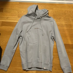 Ljusblå hoodie från Les Deux - Säljer en stilren ljusblå hoodie från Les Deux. Tröjan har en klassisk design med justerbar huva och en praktisk magficka. Perfekt för en avslappnad look.