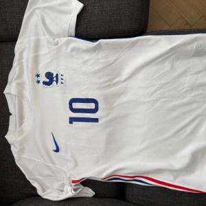 Vit frankrike tröja med nummer 10 - Säljer en vit fotbollströja från Nike med blå detaljer och nummer 10 på ryggen. Tröjan har korta ärmar och en stilren design med röd och blå rand längs sidan. Perfekt för fotbollsfans!